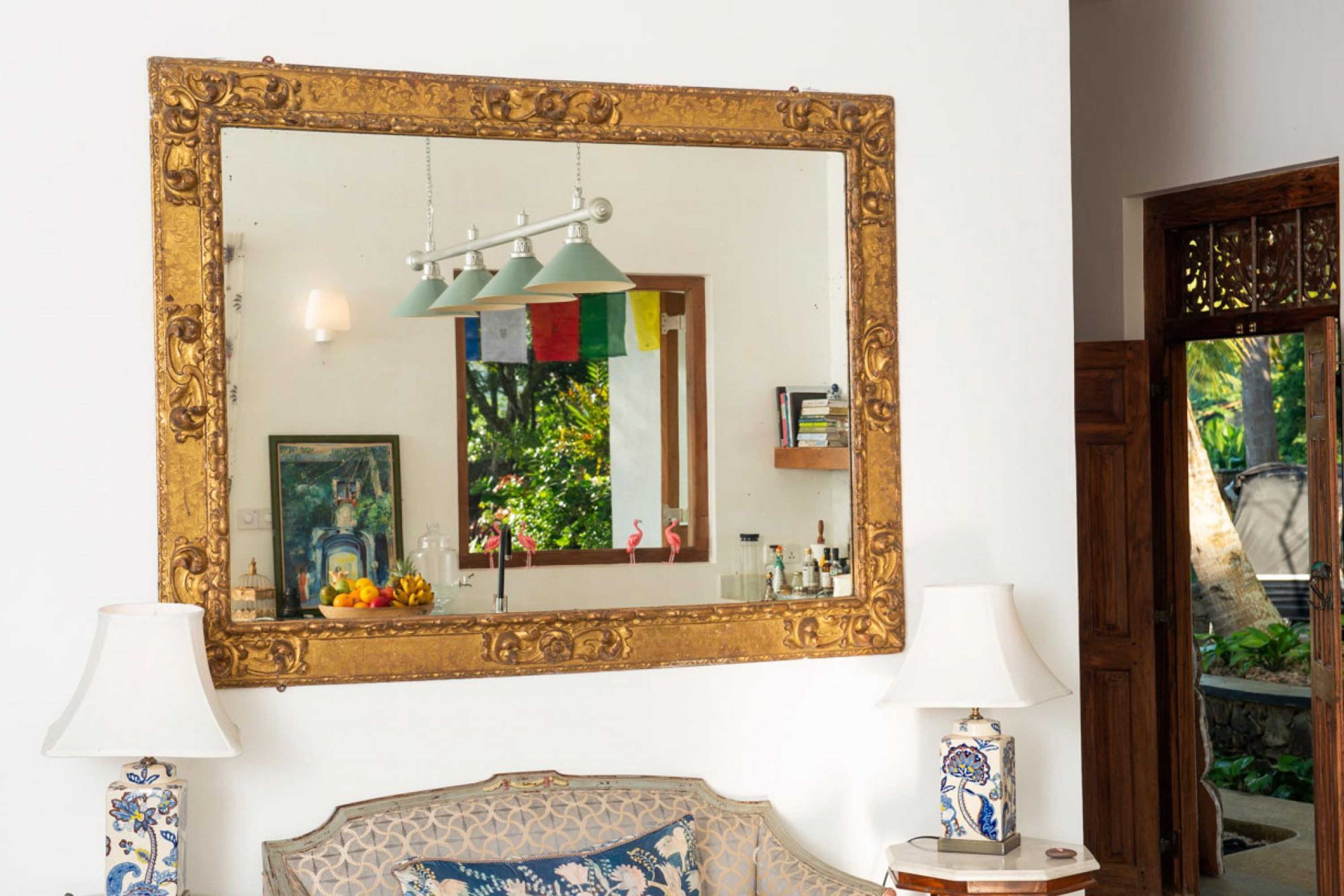Una habitación con un espejo grande y ornamentado, dos lámparas, un sofá estampado y vistas a la vegetación a través de una puerta.