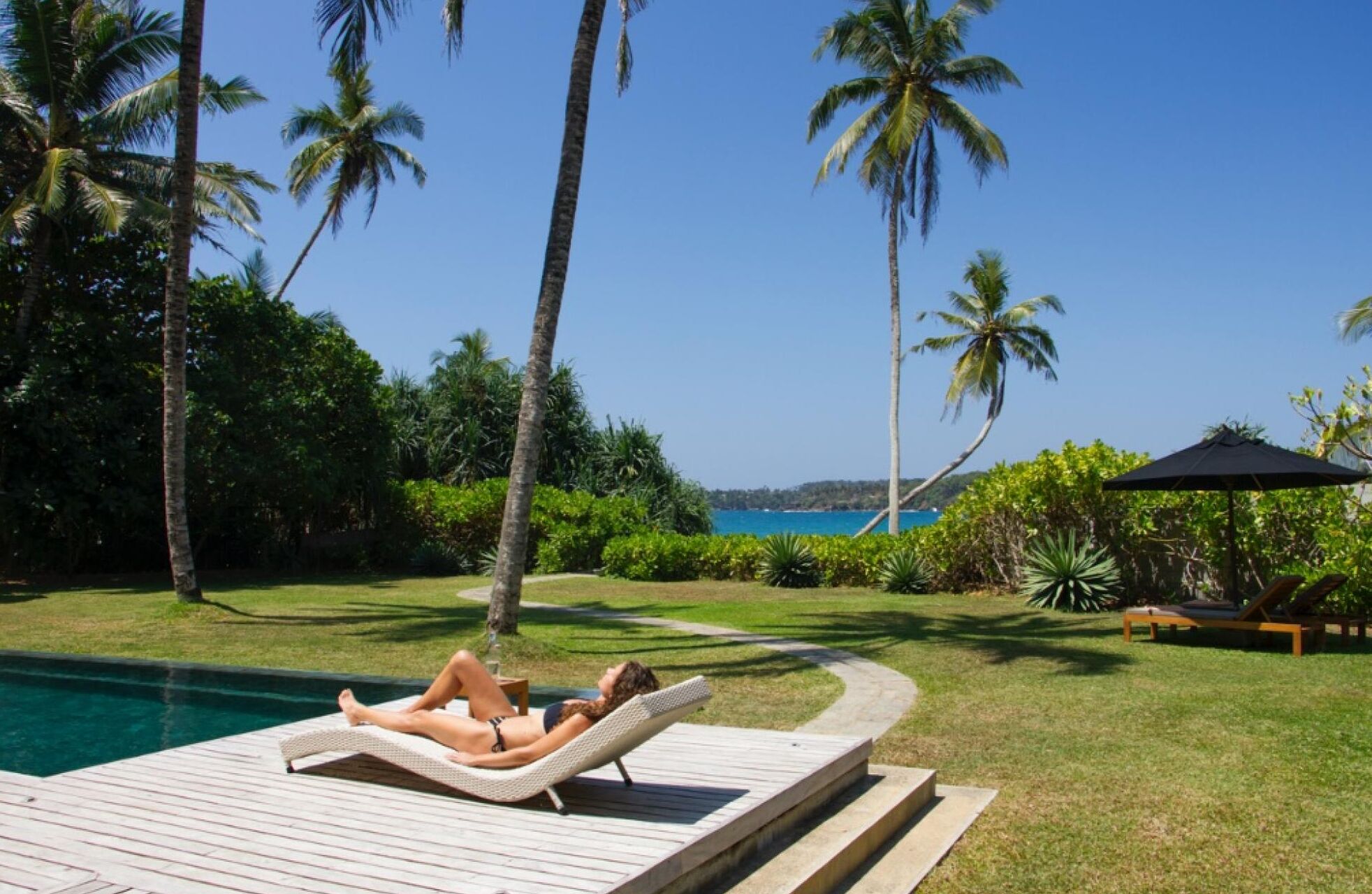 Zona junto a la piscina con sillones de descanso, palmeras y vista al océano.