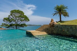 Una piscina cristalina con escalones que conducen a un muro de piedra, rodeada de árboles y vistas al mar.