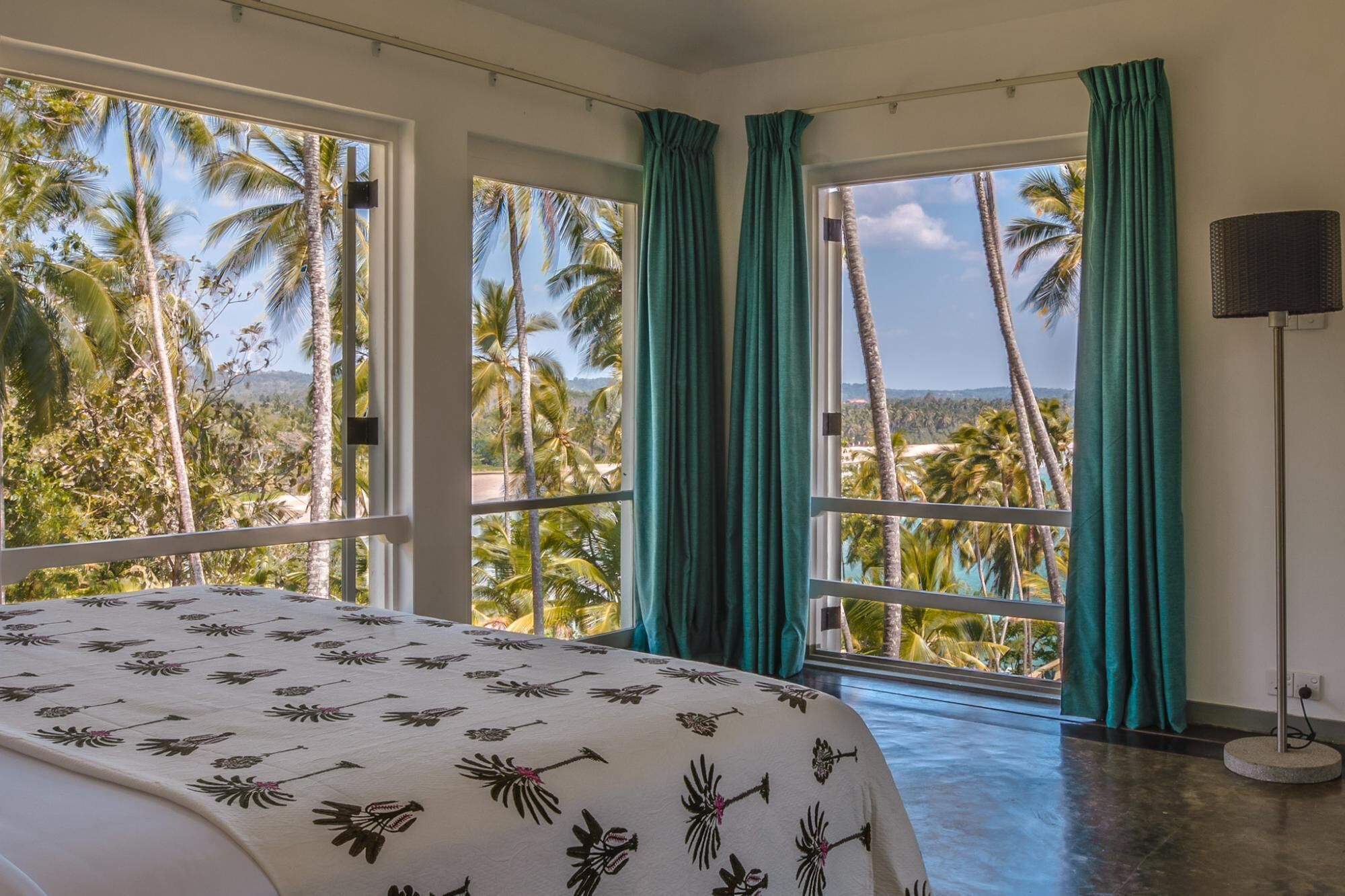 A bedroom with a bed, large windows, and a view of palm trees and a beach.