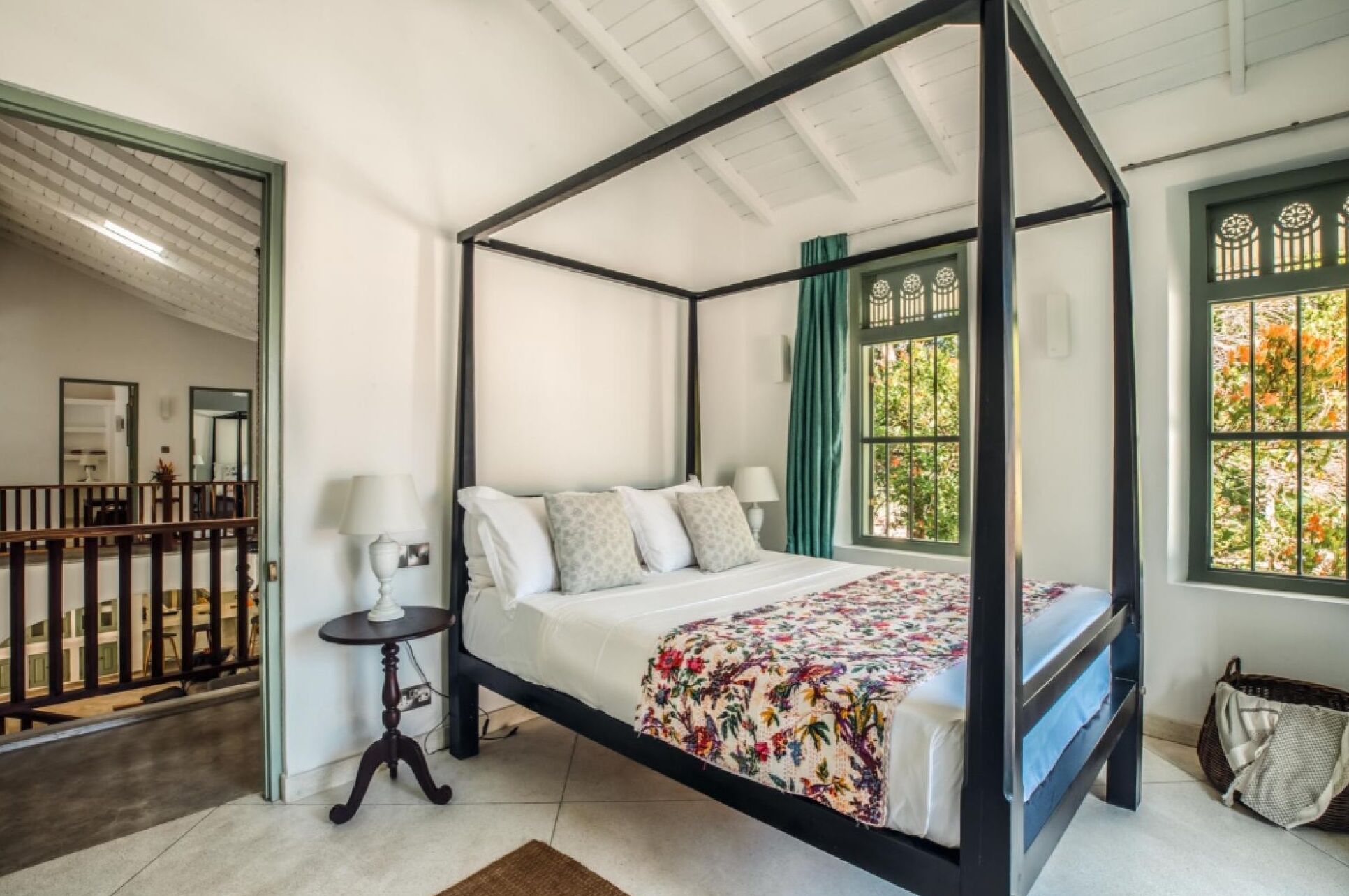A four-poster bed with a floral bedspread, a small bedside table with a lamp, and a view of the outside through a window with green curtains.