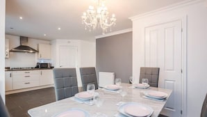 A modern dining area with a marble table set for four, grey upholstered chairs, a chandelier, and a kitchen in the background. - tms Cheerful 5bdr House! Thurrock! Free Parking! (Stanford-le-Hope)