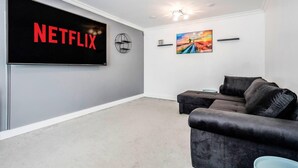 A modern living room with a large flat-screen TV displaying the Netflix logo, a cozy sectional sofa, a decorative wall piece, and a ceiling light fixture. - tms Cheerful 5bdr House! Thurrock! Free Parking! (Stanford-le-Hope)