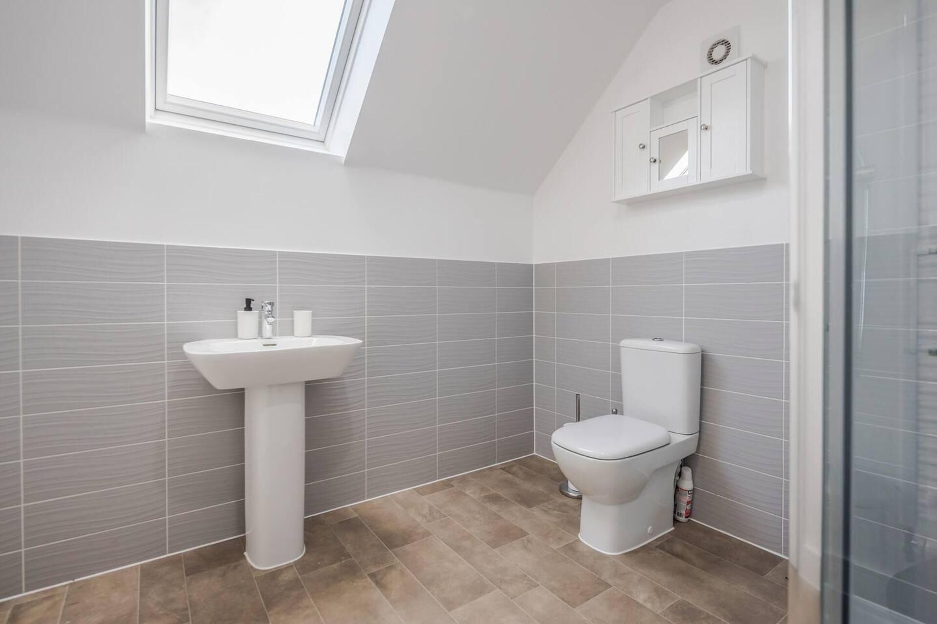 Un baño con tragaluz, lavamanos pedestal, inodoro y un mueble sobre el inodoro.