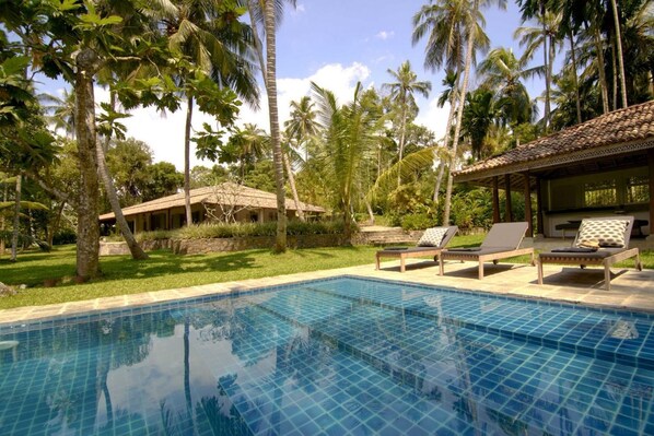 A pool area with lounge chairs, a covered seating area, and a building with a tiled roof.
