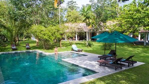 A swimming pool with lounge chairs and umbrellas, surrounded by lush greenery and a covered structure in the background. - Family House With Large Garden In A Peaceful Setting (Padinnoruwa)
