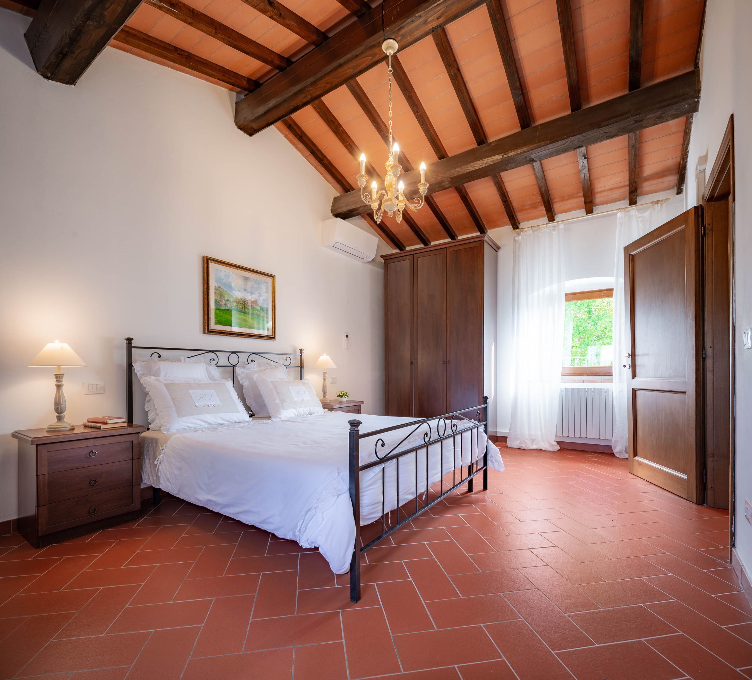 A bedroom with a bed, nightstands, a chandelier, a large wardrobe, and a window with curtains.