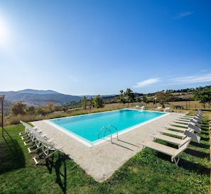 Ein rechteckiger Pool mit klarem, blauem Wasser, umgeben von einer Steindeck, und ausgestattet mit einer Leiter. Liegestühle sind rund um den Pool angeordnet, und im Hintergrund ist eine Pergola zu sehen.