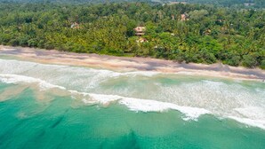 A coastal landscape with a beach, clear turquoise waters, and a backdrop of dense green forest and hills. - A Sophisticated Spacious Beach Villa With 20 Meter Water Slide (Polgahamulla)