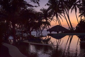 A serene pool area with palm trees and a thatched-roof structure, surrounded by lush vegetation. - A Sophisticated Spacious Beach Villa With 20 Meter Water Slide (Polgahamulla)