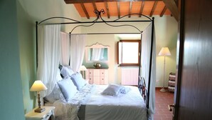A four-poster canopy bed with white linens and lace details, a bedside table with a lamp, and a view of a room with a window and a mirror. - Iris apartment (Città metropolitana di Firenze)