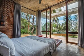 A bedroom with a large bed, a brick wall, and a view of palm trees through a sliding glass door.