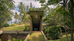 A small, enclosed structure with a brick base and a glass door, surrounded by lush greenery and palm trees, overlooking a body of water. - Sustainably Designed Villa Overlooking Indian Ocean (Tangalle)