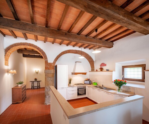 Uma cozinha com armários brancos, uma ilha central, um arco de tijolos e teto de madeira rústica.