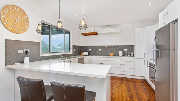 Une cuisine moderne dotée d’un îlot central, d’une horloge en bois, de luminaires suspendus et d’une grande fenêtre.