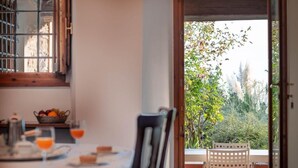 Un comedor con una mesa lista para dos personas, una silla y vista al jardín a través de una puerta de cristal.