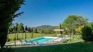 Una piscina con sillas de descanso, rodeada de césped y árboles, con vistas a colinas al fondo.