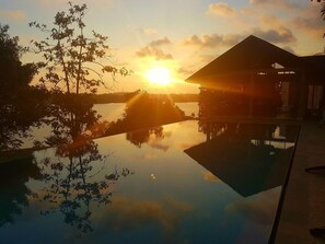 Una puesta de sol serena sobre una masa de agua tranquila, con su reflejo, rodeada de árboles y una estructura de edificio.
