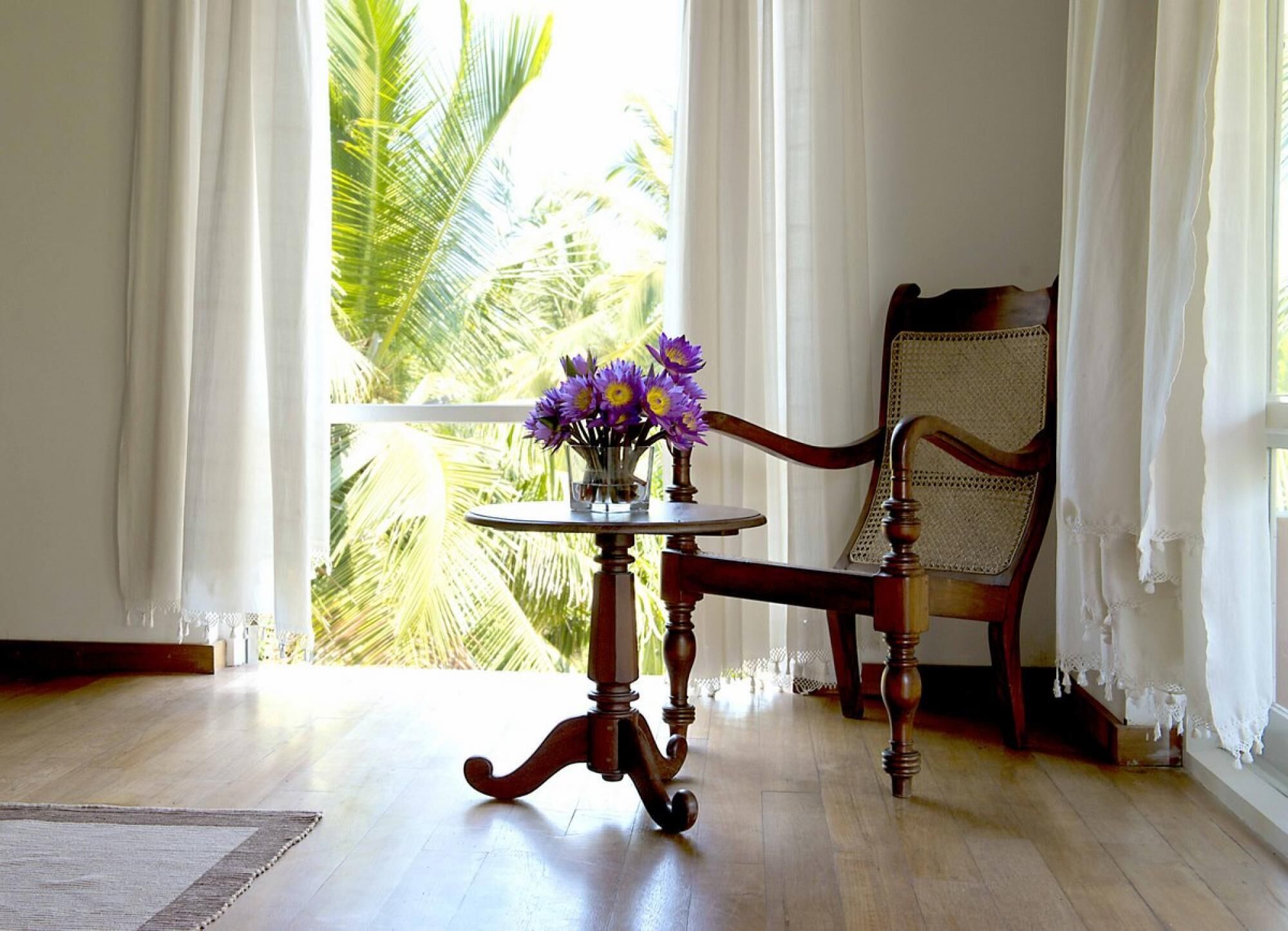 A room with a wooden table and chair, a vase with flowers, and a view of palm trees through sheer curtains.