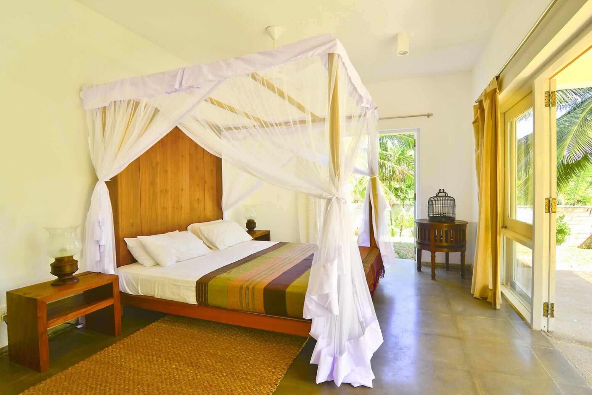 A bedroom with a canopy bed, a wooden headboard, a bedside table with a lamp, a woven mat, and a view of greenery outside.