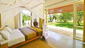 Un dormitorio con cama, mesita de noche, jaula de pájaros y vista a un jardín a través de un ventanal.