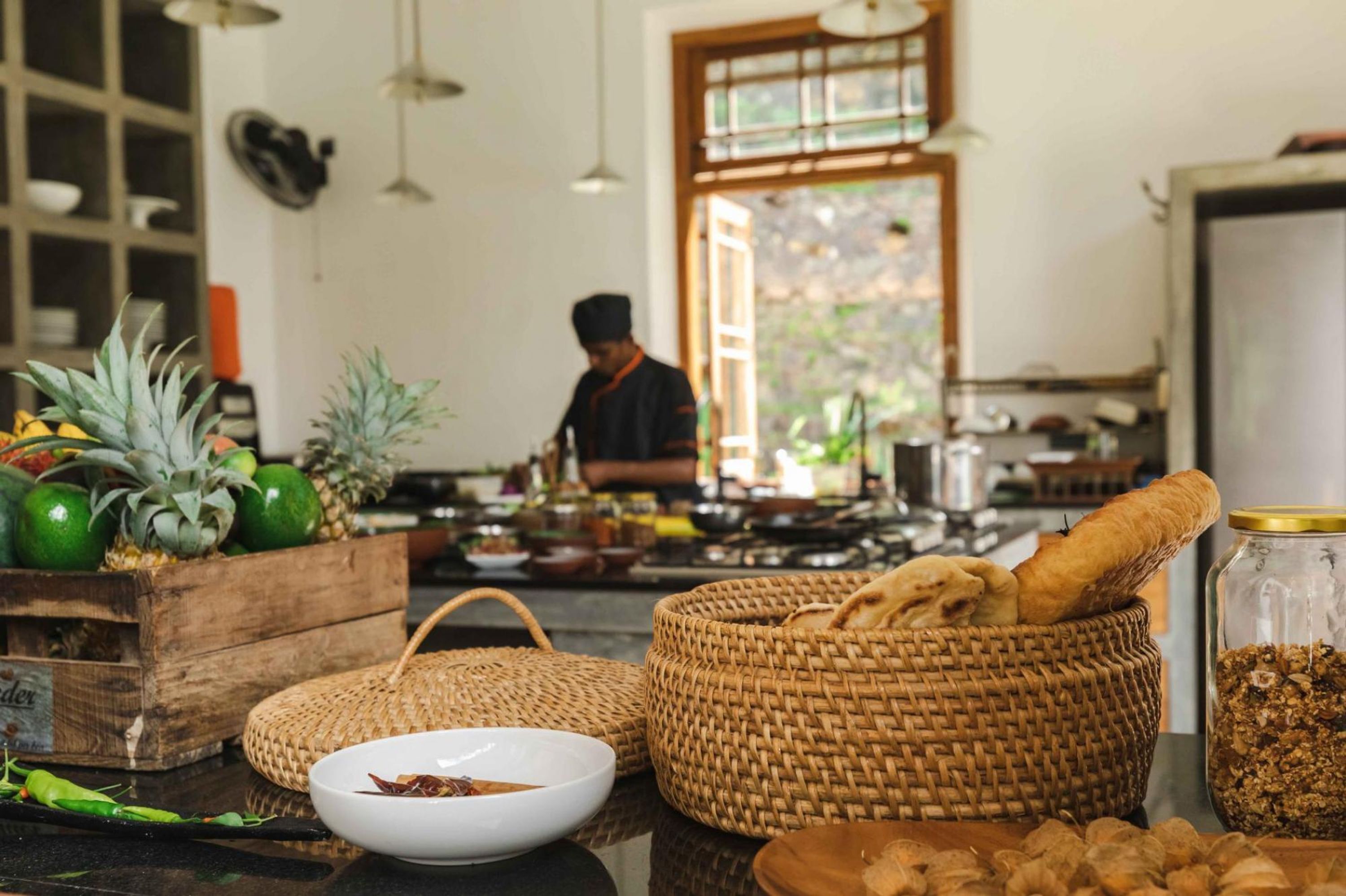 A kitchen with a wooden table displaying fruits, bread, and a bowl, and a person cooking in the background.