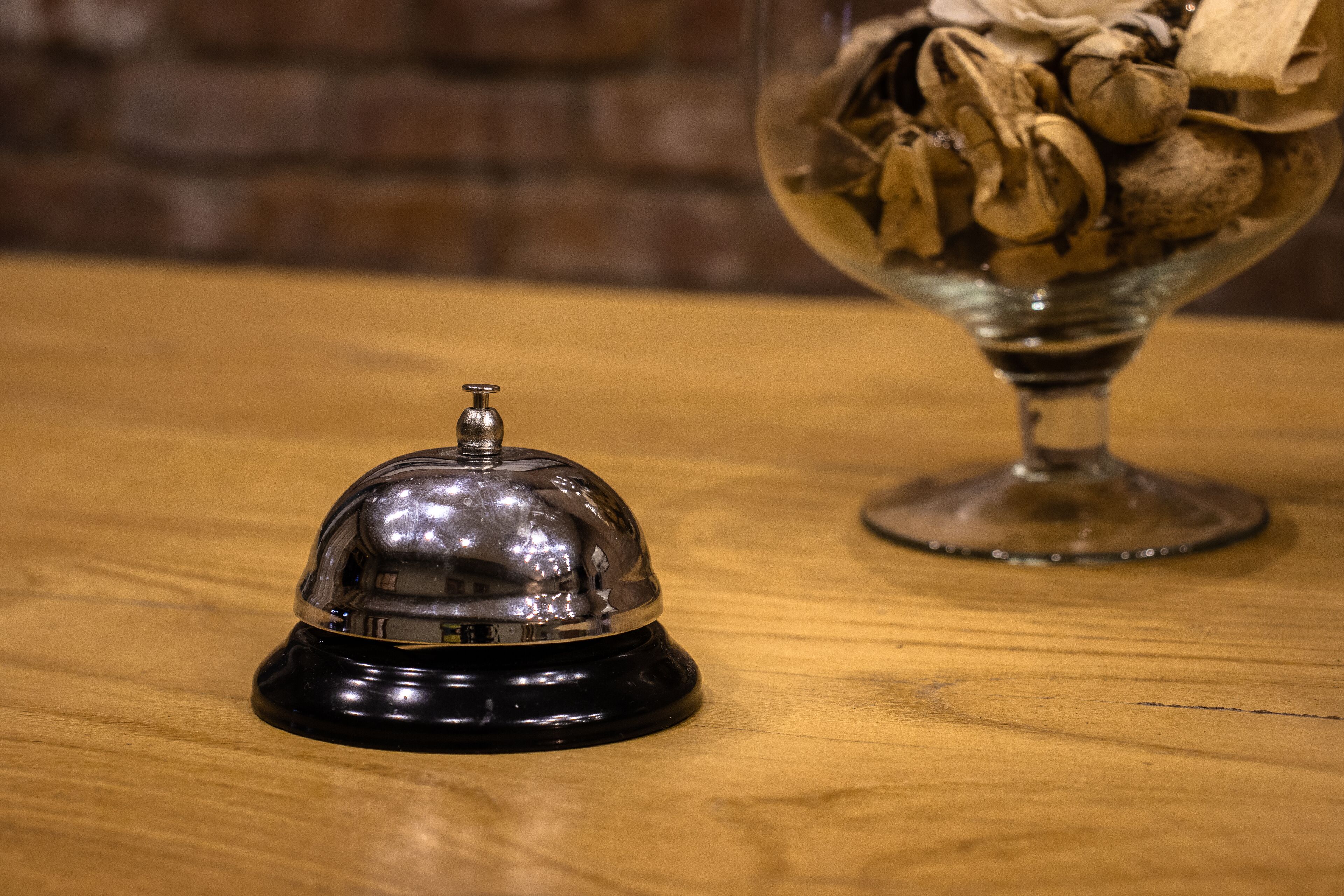 A black bell on a wooden surface, a glass bowl with nuts, and a brick wall in the background.