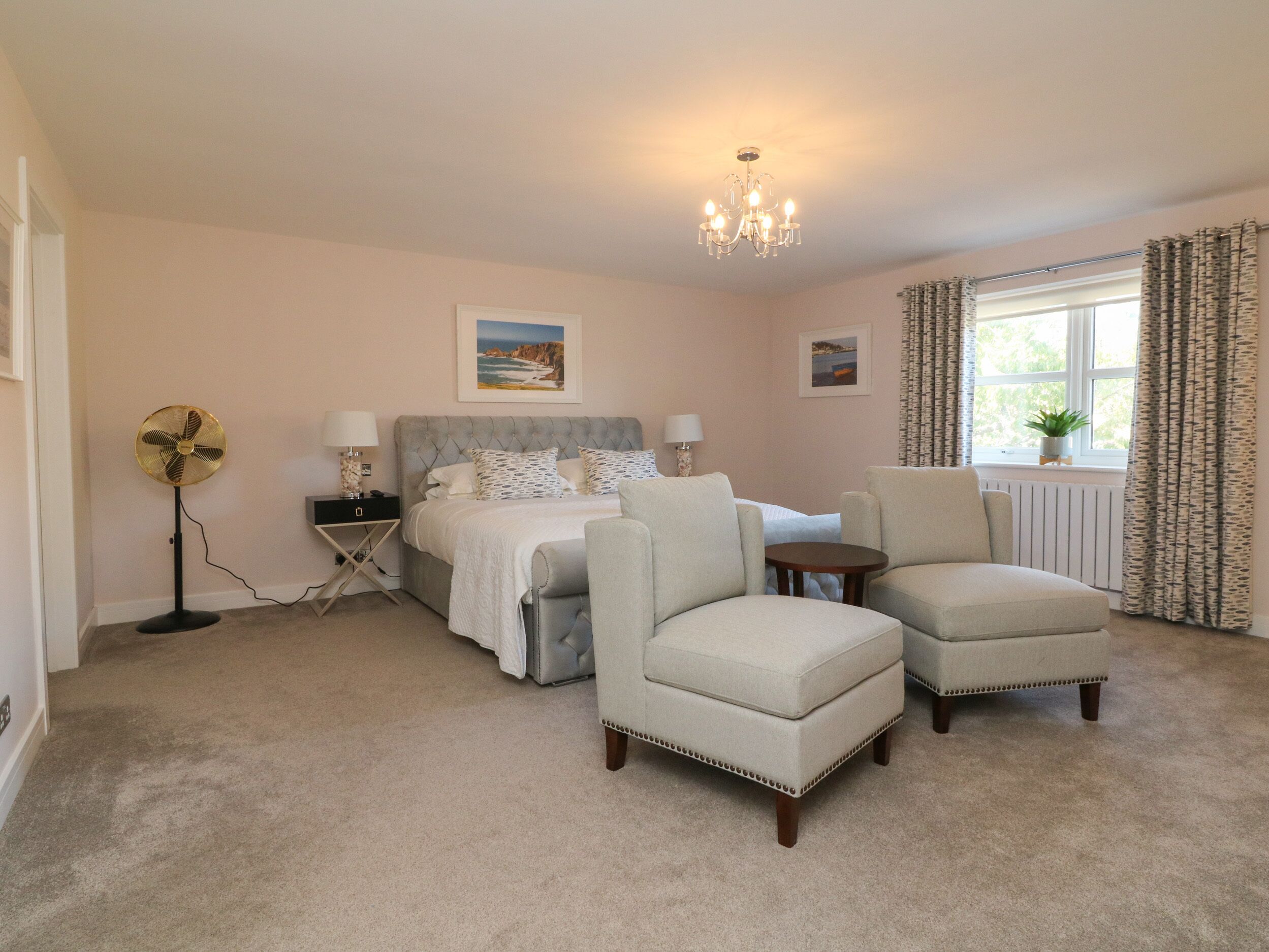 A bedroom with a bed, two armchairs, a standing fan, a bedside table, and a window with curtains.