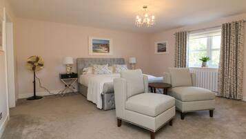 A bedroom with a bed, two armchairs, a standing fan, a bedside table, and a window with curtains.