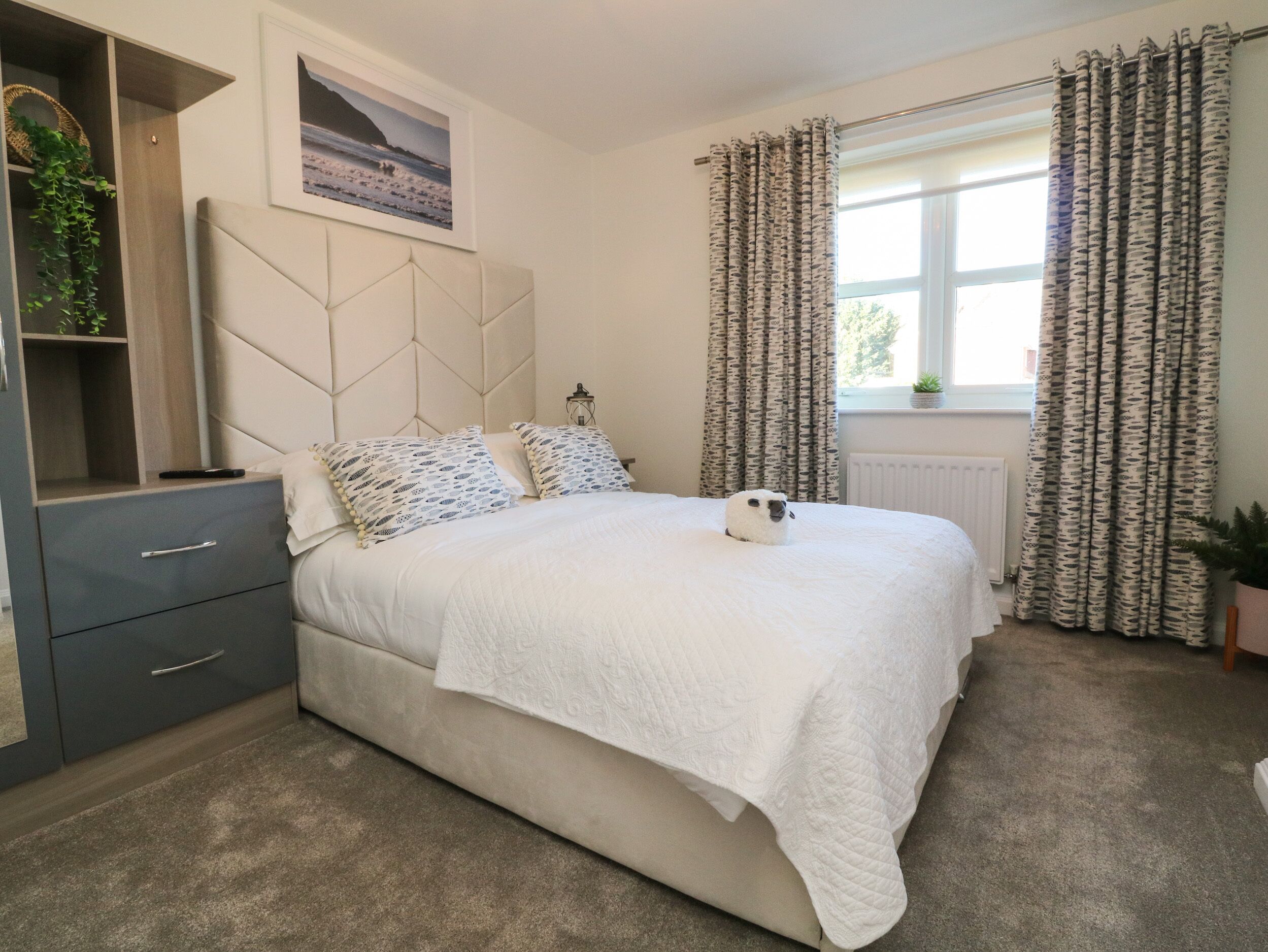 Une chambre à coucher comprenant un lit, une table de chevet, une commode, une tête de lit, une fenêtre avec des rideaux et une étagère murale agrémentée d’une plante.