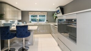 A modern kitchen with a central island, bar stools, a built-in oven, and a window with a view of greenery. - 4 Woodland Park (Bideford)