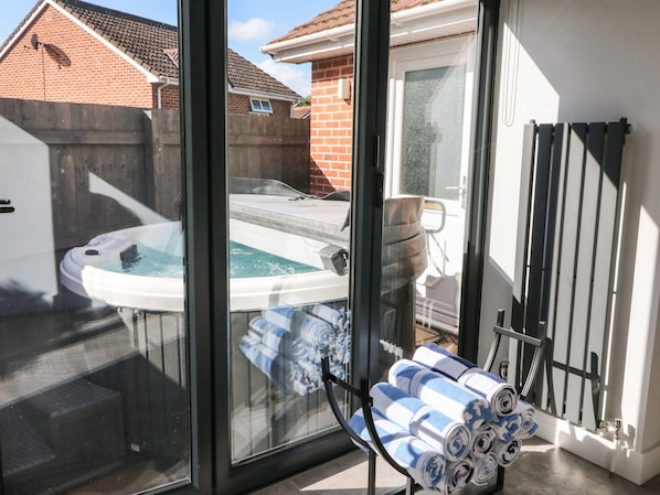 A glass-enclosed patio with a hot tub, a towel rack with towels, and a brick house in the background.