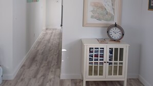 A hallway with a sign reading "BEACH HOUSE," a framed artwork of a bird, a clock, and a small cabinet with drawers.