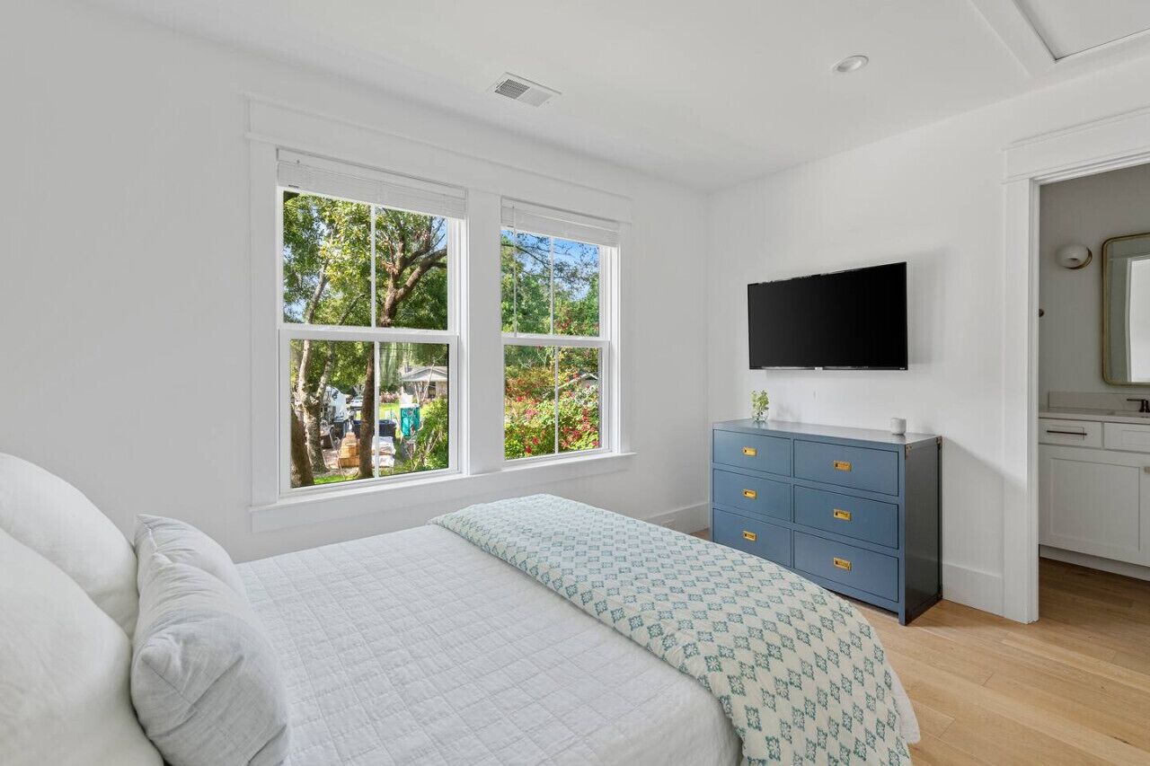 A bedroom with a bed, a dresser, a TV, and a window with a view of trees and a garden.