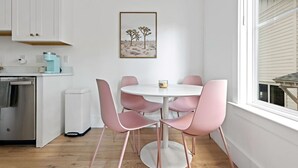 A modern kitchen with white cabinets, a round table with pink chairs, and a desert landscape painting on the wall.
