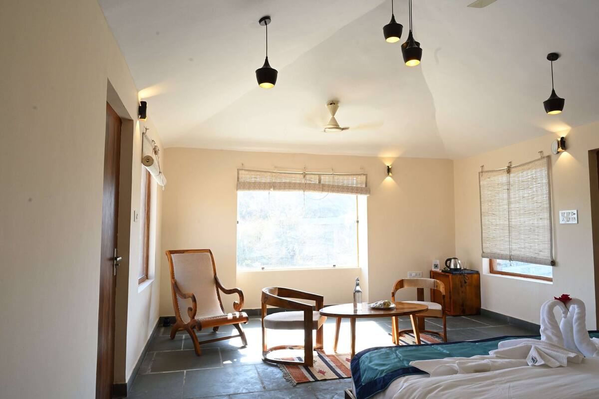 A room with a bed, two chairs, a small table, a window with blinds, a ceiling fan, and pendant lights.