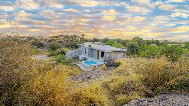 Una casa de piedra con piscina, rodeada de hierba seca y árboles, bajo un cielo parcialmente nublado.