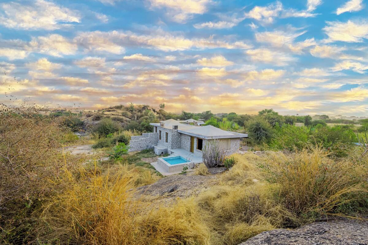 A stone house with a pool, surrounded by dry grass and trees, under a partly cloudy sky.
