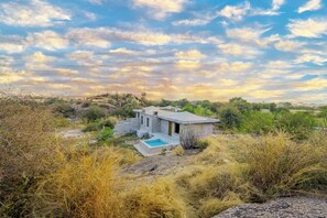 A stone house with a pool, surrounded by dry grass and trees, under a partly cloudy sky. - Tree of Life of Leopard Caves, Jawai (Bali)