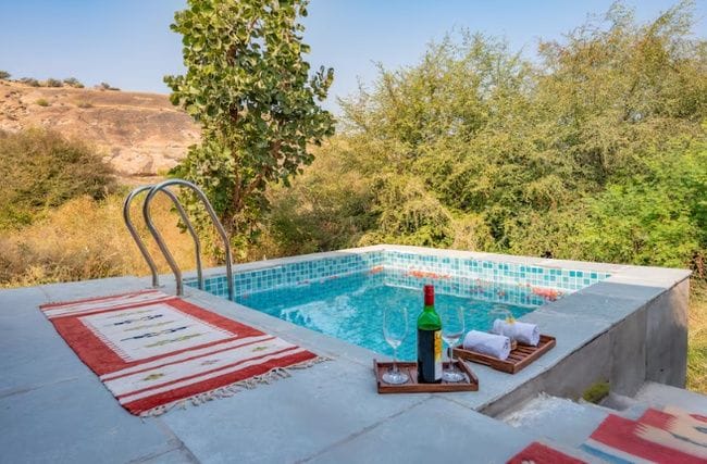 A rectangular outdoor swimming pool with a tiled edge, surrounded by greenery and a clear sky.