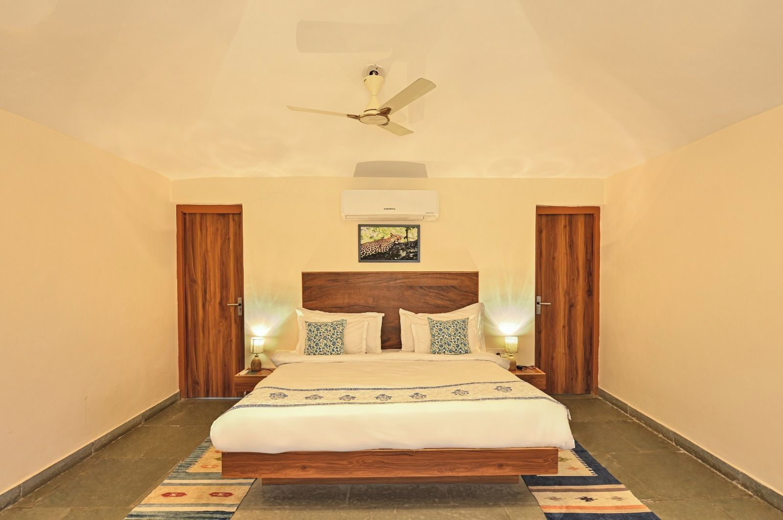 A bedroom with a bed, wooden headboard, two nightstands, a ceiling fan, and a wall-mounted air conditioner.