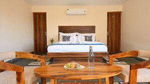 Habitación de hotel con cama, sillas de madera, una mesa con fruta y una botella, y un equipo de aire acondicionado en la pared.