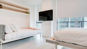 A modern bedroom with a bed, a sofa bed, a mounted TV, and a large window with a city view. - Modern & newly built studio Pool|Gym|Restaurant| (Miami)