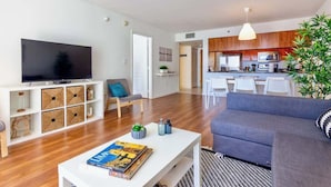 A modern living room with a flat-screen TV, a grey sofa, and a white coffee table with a book. A kitchen with wooden cabinets and white appliances is visible in the background. - Ocean & City Views Lux Miami Brickell | Sleeps 5 (Miami)