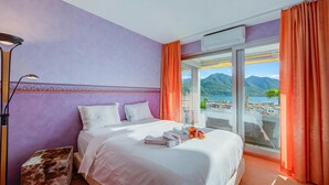 A hotel room with a bed, a nightstand, a lamp, and a view of mountains and water. - Panorama maggiore apartment (Locarno)