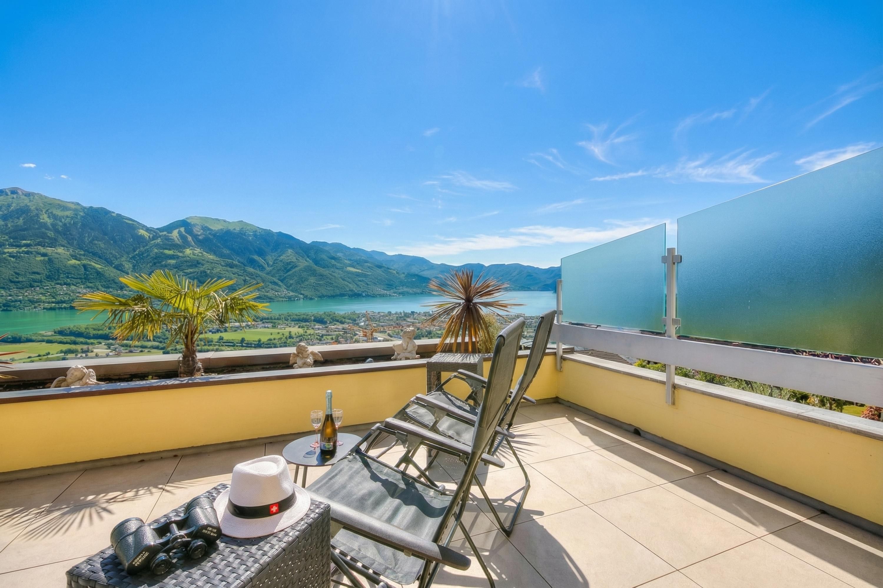 A balcony with a view of mountains and a lake, a table with a bottle, binoculars, and a hat.