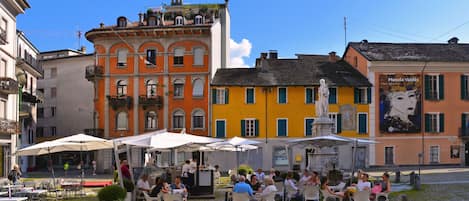 An outdoor cafe with tables and chairs, surrounded by colorful buildings and a statue.