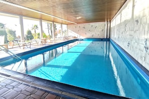 Piscina interior com água cristalina, teto de madeira, paredes revestidas de azulejos e janelas iluminadas pelo sol.
