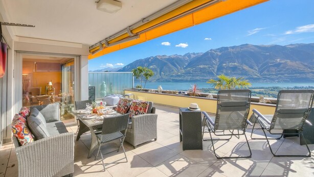 A balcony with wicker furniture, a table, and chairs, overlooking a mountainous landscape and a body of water. - Panorama maggiore apartment (Locarno)
