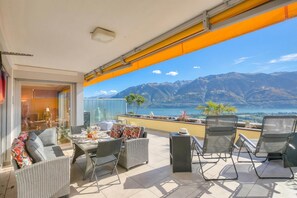 Un balcone con mobili in vimini, un tavolo e sedie, con vista su un paesaggio montuoso e uno specchio d'acqua.